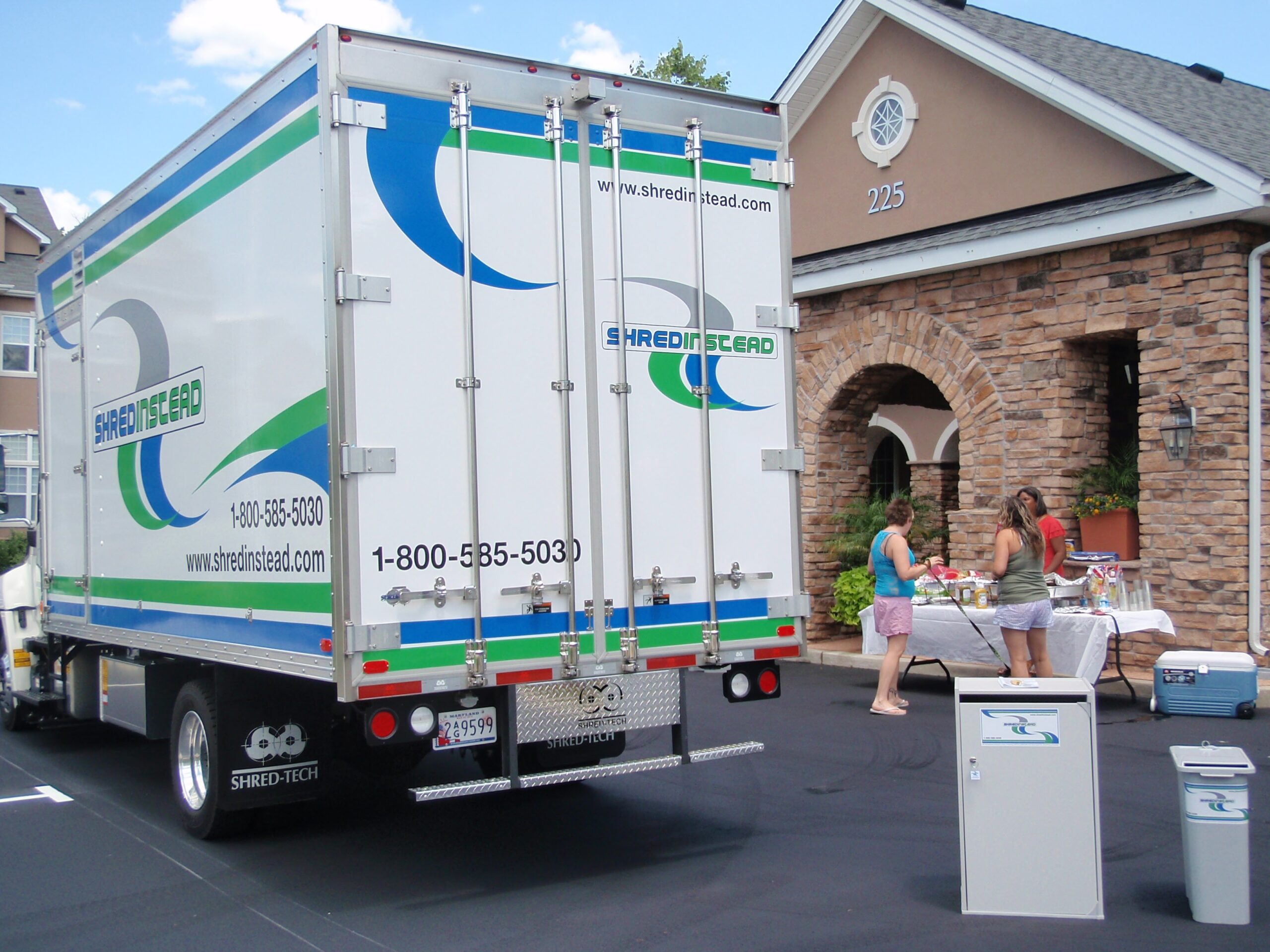 The image shows a Shred Instead mobile shredding truck parked outside a residential-style building with stone accents and the address number “225” displayed near the entrance. In the foreground, secure document collection bins with the company’s branding are placed on the pavement. Nearby, a few people stand at a table set up outside, suggesting a community shredding event or document drop-off day.