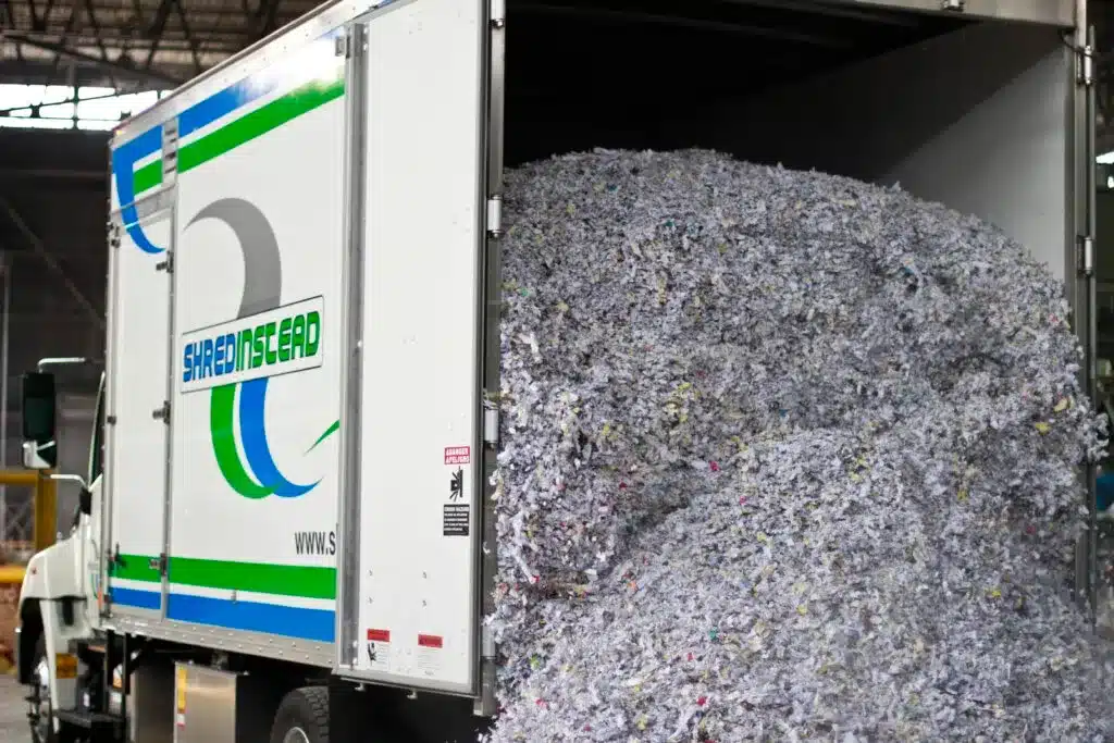 Photograph of a white box truck from "SHREDINSTEAD" with its rear door open, revealing a large pile of shredded paper. The paper is mostly white and grey with colorful flecks. The truck's side has a blue and green logo with the company name.