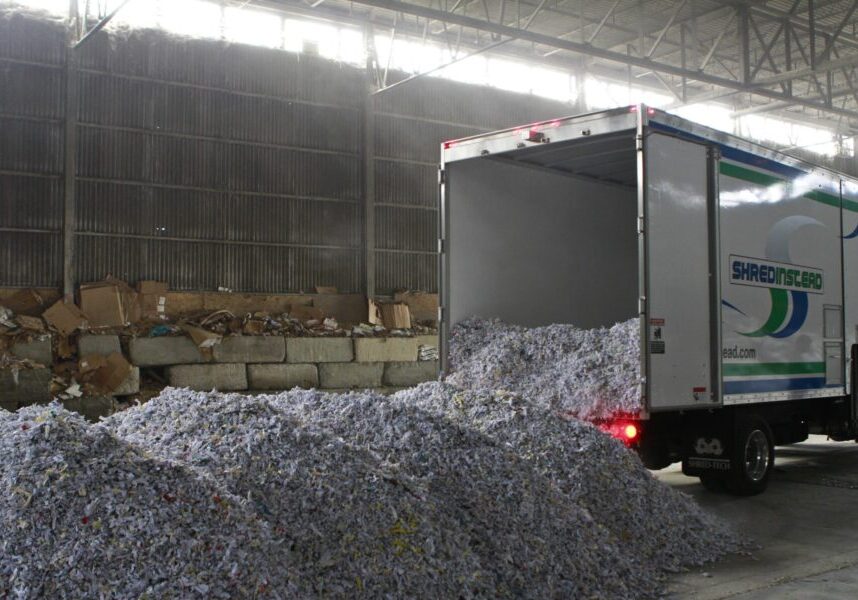 The image shows the rear of a Shred Instead mobile shredding truck inside a large industrial warehouse. The back door of the truck is open, and a large pile of shredded paper has been unloaded onto the concrete floor behind it.