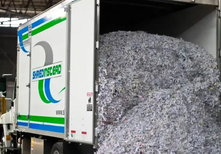 Photograph of a white box truck from "SHREDINSTEAD" with its rear door open, revealing a large pile of shredded paper. The paper is mostly white and grey with colorful flecks. The truck's side has a blue and green logo with the company name.